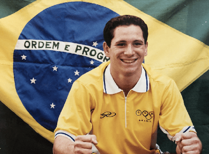 Tiago Camilo jovem sentado com camisa amarela e bandeira do Brasil ao fundo